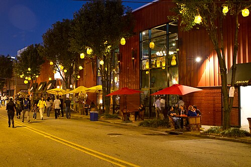 A red corrugated metal building at night during the Second Friday Art Stroll, string lights in the trees and people seated under umbrellas on the sidewalk.