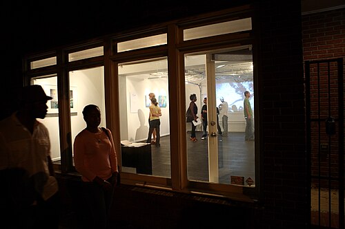 A lit gallery storefront at night during the Second Friday Art Stroll, framed pieces on the walls visible through the window and two visitors on the sidewalk outside.