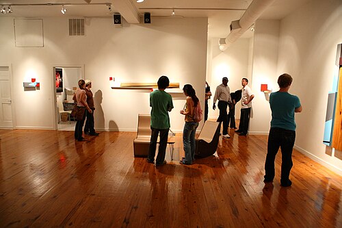 The inside of a bare loft gallery during the Second Friday Art Stroll, several visitors standing near a few small pieces mounted on the walls.