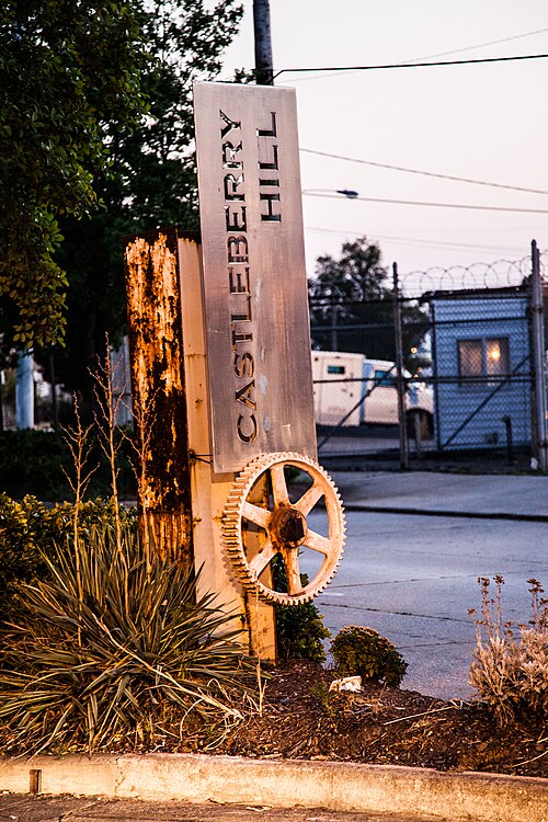 A rusted metal sign reading Castleberry Hill with an old gear wheel bolted to its base, next to a chain link fence at dusk.