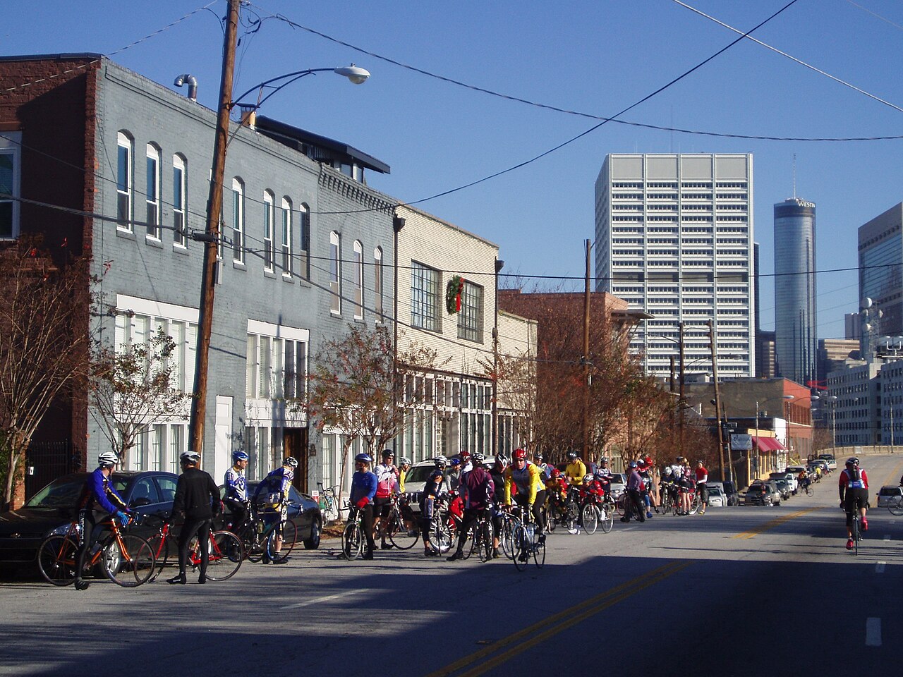 A street of brick and painted loft buildings with cyclists gathered outside them, the downtown Atlanta skyline rising at the far end on a clear day.