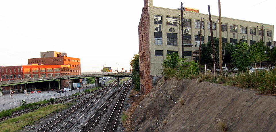 A view from a bridge over freight tracks, an old brick mill building beyond them with faded painted lettering across its upper floors, and another industrial building further down the line.