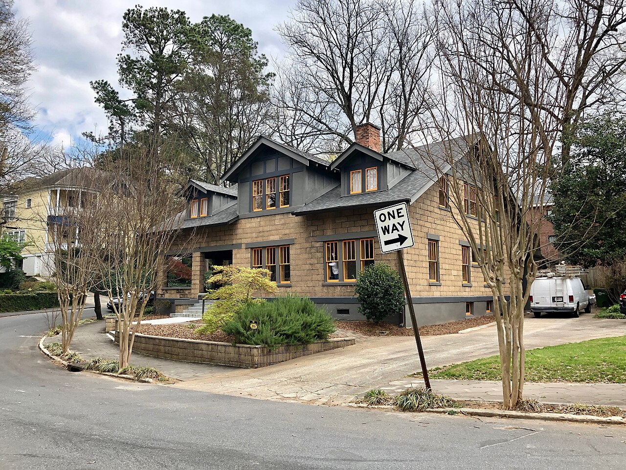 The bend in Druid Circle, a shingled house set above a low wall with crepe myrtles along the kerb and a one way sign at the junction.