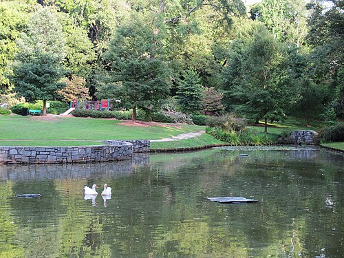 The pond in the neighborhood park, geese on the water, a stone retaining wall along the near bank and a playground on the grass behind it.