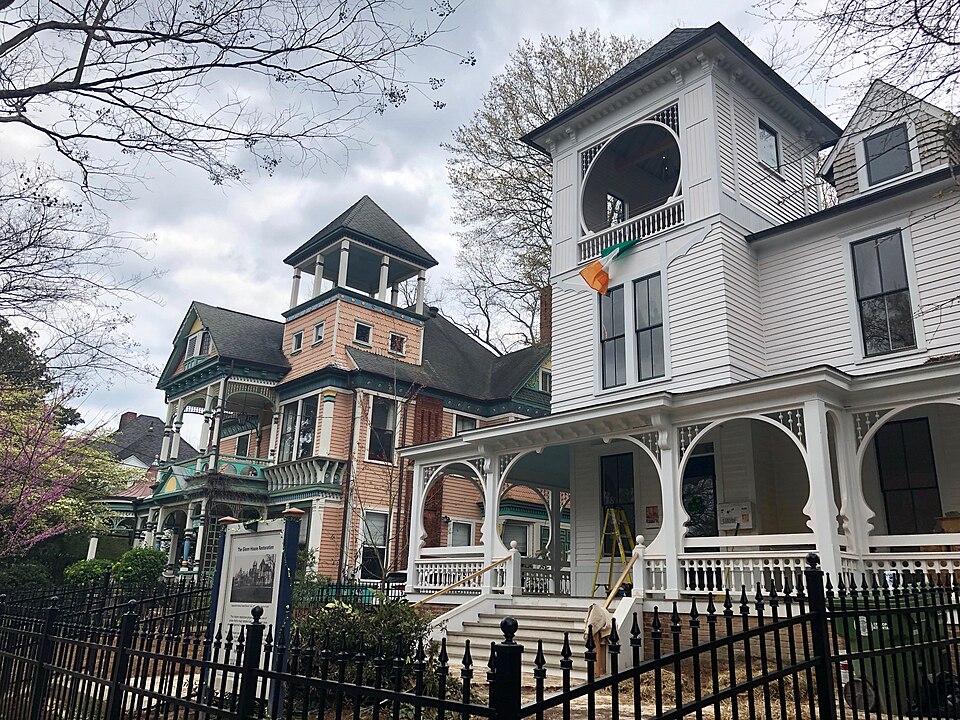 Two Queen Anne houses standing side by side behind a spiked iron fence, the white one with an open arched loggia at the top and a salmon coloured one next to it under a square cupola.