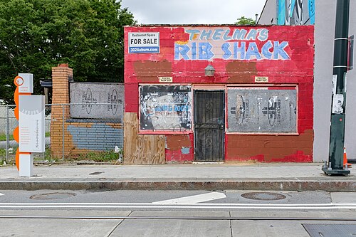 The former Thelma's Rib Shack at 302 Auburn Avenue, a painted cinder block storefront boarded up with a for sale sign on the wall and streetcar rails in the road in front of it.