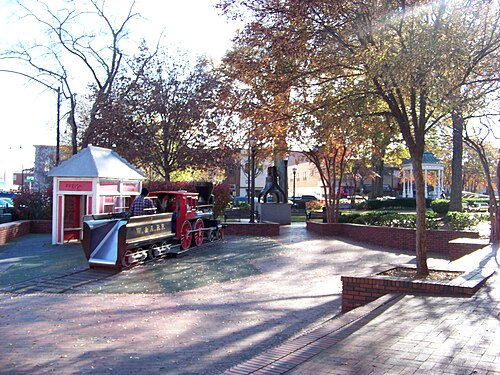 A small scale steam locomotive on a short length of track in the park, a red trackside booth beside it and a statue among the trees beyond.