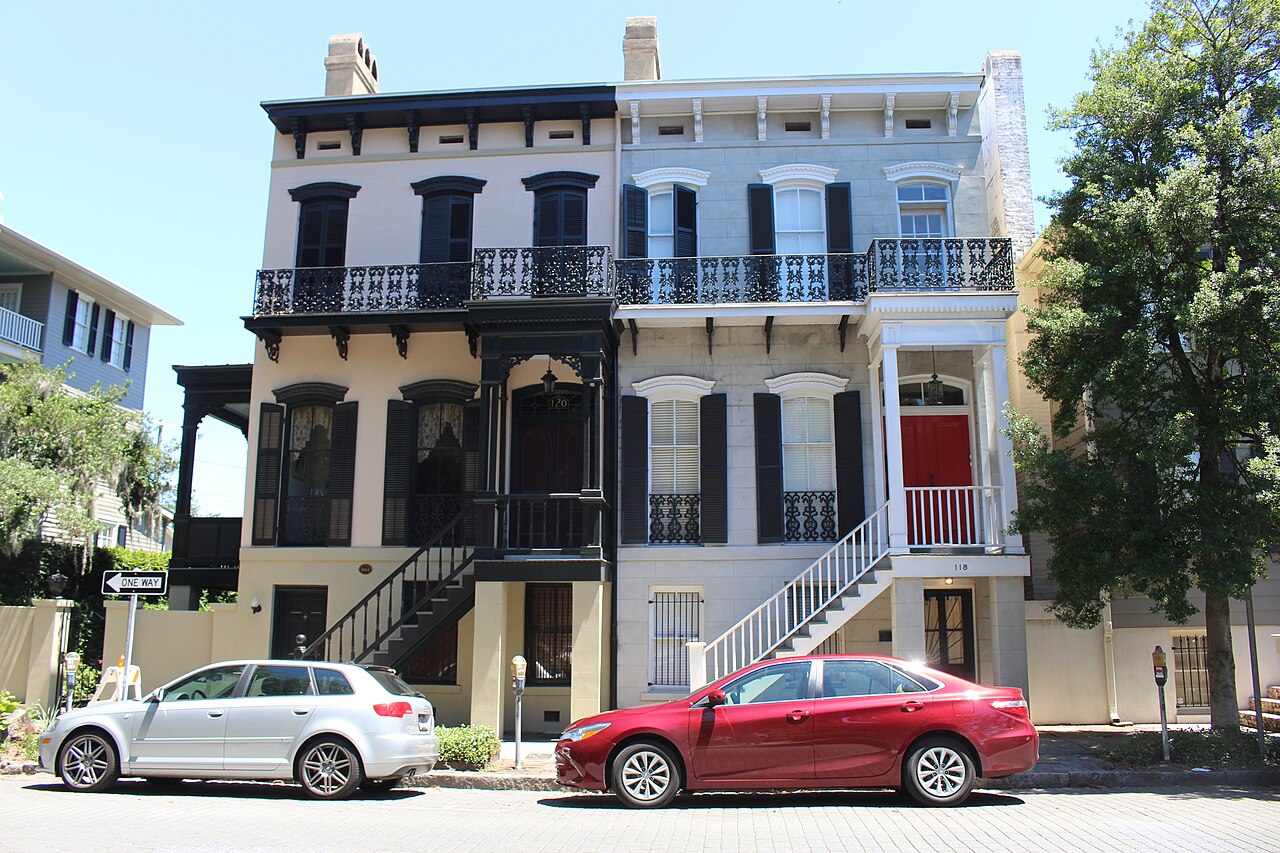 A pair of attached Italianate townhouses in cream stucco and pale stone, each with a two story iron balcony and a red or dark painted front door reached by a straight stoop.