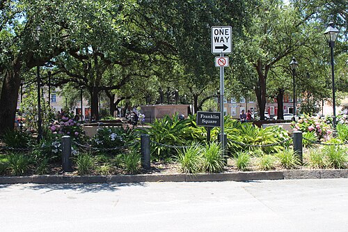 Franklin Square's monument and garden beds under a canopy of live oaks, with a one way street sign, flowering shrubs and a row of townhouses beyond the trees.