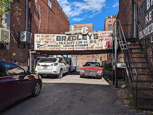 A rear brick alley between two warehouse buildings, a large hand painted metal sign for a locksmith spanning overhead and an exterior iron stair climbing the wall.
