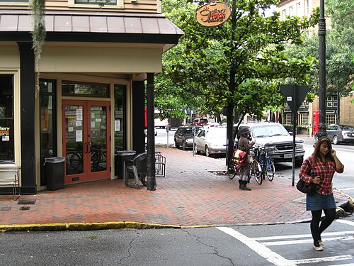 A corner storefront on Bull Street under a standing seam metal awning, brick paved sidewalk, a magnolia tree and parked bicycles at the curb.