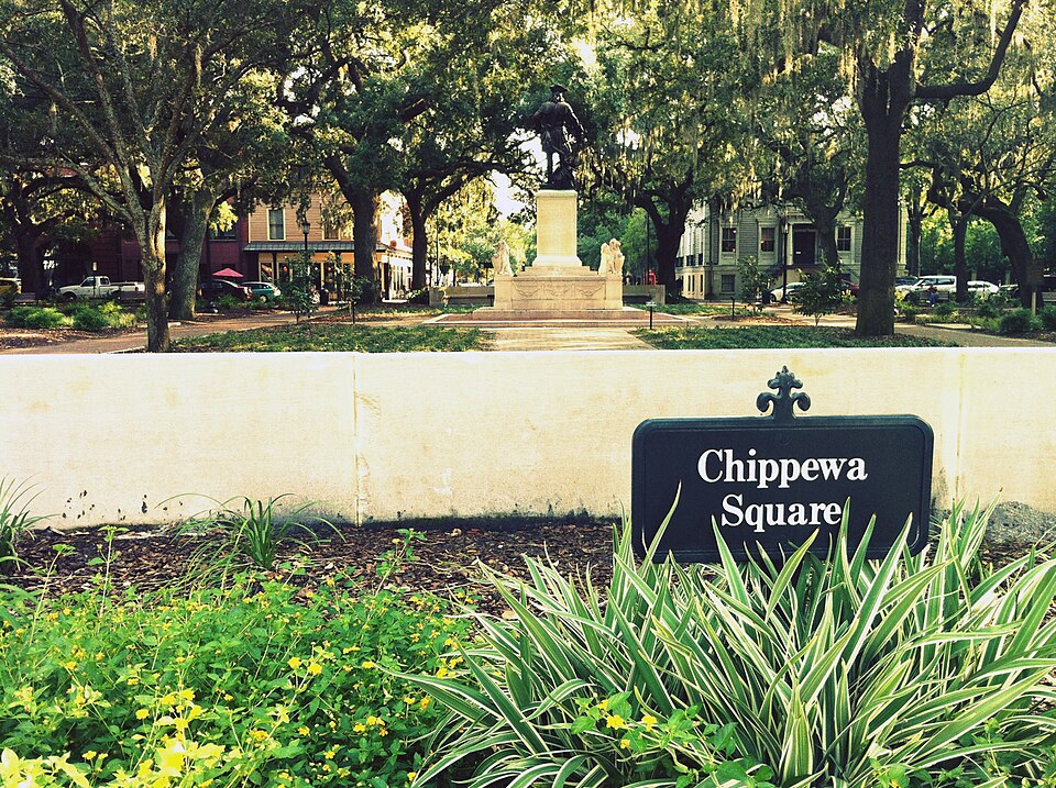 Chippewa Square's own sign in the foreground, with the James Oglethorpe Monument under the oak canopy behind it and townhouses along the square's far side.