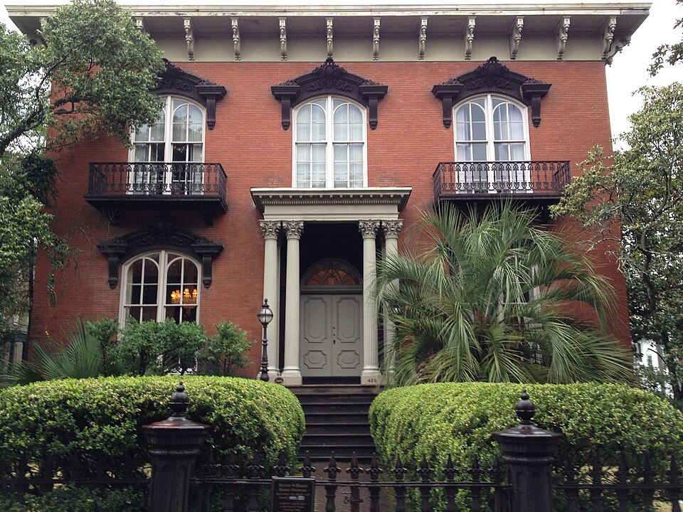 The Mercer Williams House's red brick Italianate facade straight on, with its iron balconies, columned entry porch and the museum sign at the front gate.