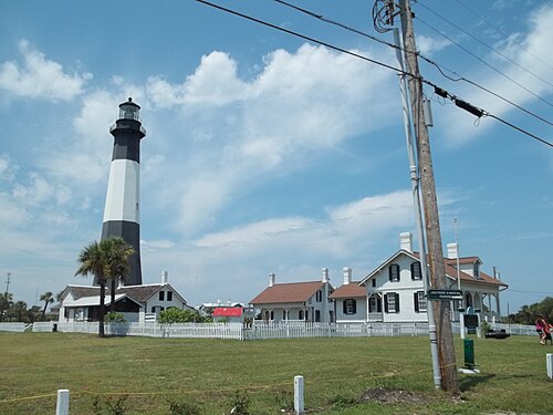 The black and white Tybee Island Lighthouse behind a white picket fence, with the lightkeeper's house and several smaller outbuildings on the lawn beside it.