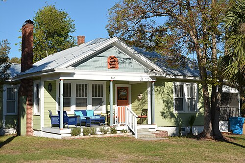 A single storey light green cottage numbered 802, a brick chimney, and a small covered porch with patio furniture.