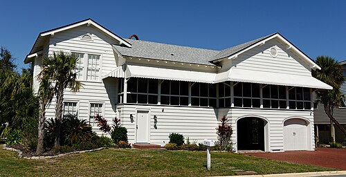 A two storey white cottage raised over an enclosed ground floor with a garage door, palms in the yard and a screened porch above.