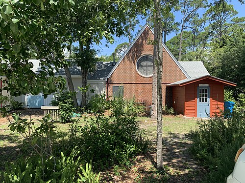 A small brick church with a round stained glass window in its gable end, beside a red outbuilding with a blue door.