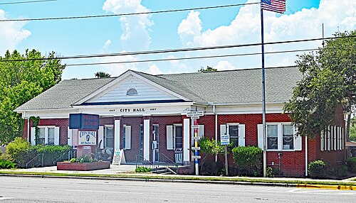 A single storey red brick municipal building with white columns, 1887 and 1987 carved into its pediment, and a flagpole out front.
