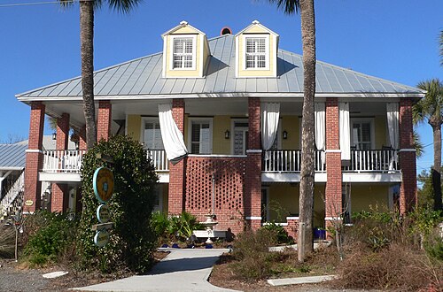 A two storey beach house with brick ground floor piers, double wraparound porches, and palm trees along the front walk.