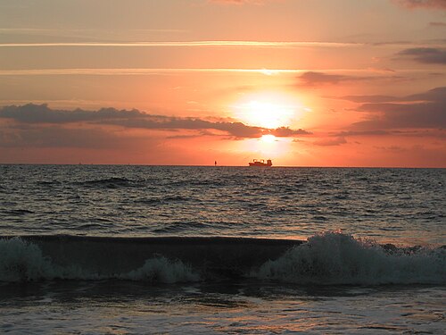 A wave breaking in the foreground at sunrise, a cargo ship silhouetted against the sun on the horizon.