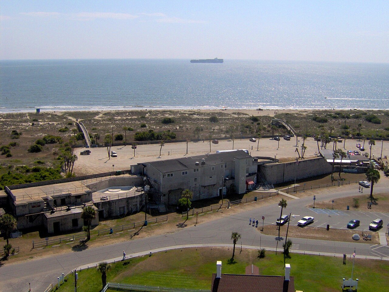 The view east from atop the lighthouse over Fort Screven's grounds and a wooden dune crossing to the beach, a cargo ship on the horizon.