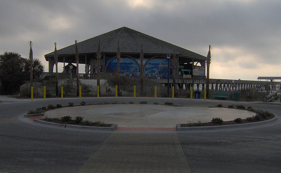 The octagonal pavilion with arched openings around it under a single peaked roof and a mural under two of the arches, the wooden pier running past it toward the ocean, seen across a plaza with a traffic circle.