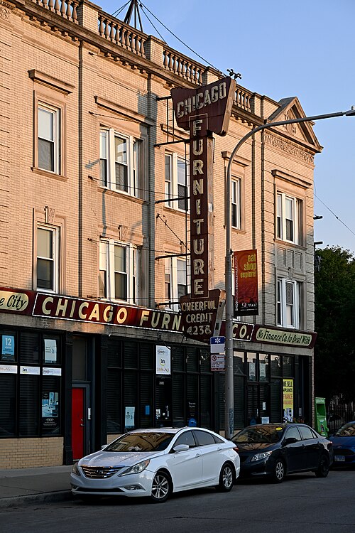 A tan brick commercial building on Cottage Grove Avenue, a tall vertical neon furniture store sign over the storefront and cars parked at the curb.