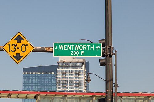 A green South Wentworth Avenue street sign under a height clearance marker, apartment towers in the distance behind.