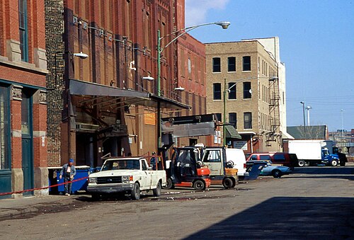 A brick warehouse loading dock under a metal canopy, a pickup truck and a forklift at the curb, a worker beside the dock.