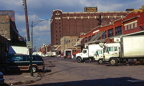 A tall brick cold storage warehouse with a large painted Fulton Market sign near its roofline, delivery trucks parked below.