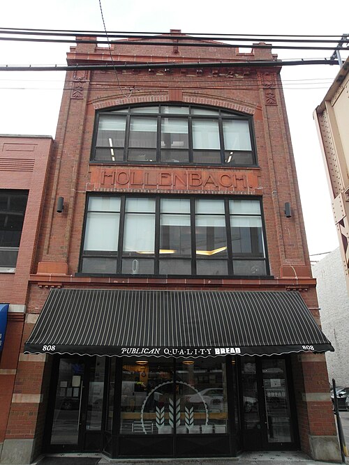 A narrow three storey red brick building with a carved terra cotta name plate reading Hollenbach, a striped awning over a bakery storefront at street level.