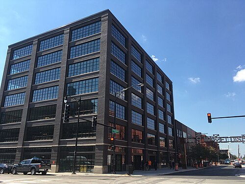 A dark brick and glass six storey office building on a corner, the Fulton Market District gateway sign visible down the block.