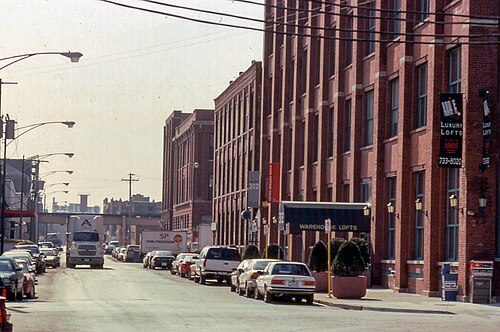 A red brick warehouse with a Warehouse Lofts banner reading Luxury Lofts Since 1985, an elevated rail bridge down the street, parked delivery trucks.