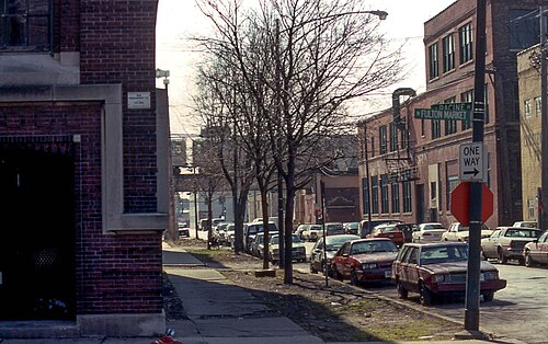 Street signs reading Racine and Fulton Market at a one way intersection, bare trees, parked cars and low brick buildings on the corner.
