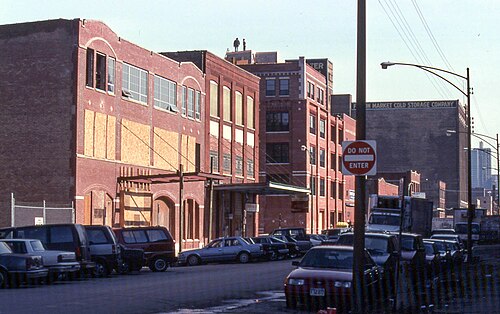 A row of brick warehouses with boarded upper windows and a loading canopy, a faded painted wall sign reading Fulton Market Cold Storage Company on a taller building behind.