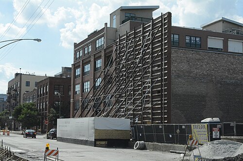 A brick warehouse mid renovation with steel scaffolding braced against one wall, converted loft buildings alongside, a demolition contractor's sign on the fence.