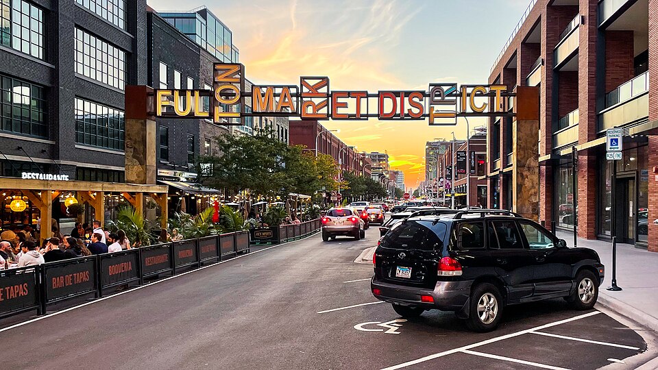 The Fulton Market District gateway sign over the street at dusk, restaurant tables and diners along the sidewalk beneath it.