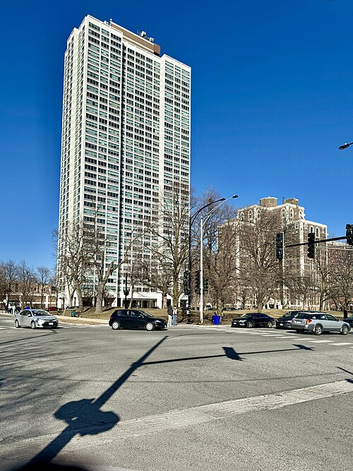A white modern high rise apartment tower on 56th Street in Indian Village, seen across a wide intersection from older brick buildings.
