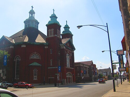 A red brick church with three copper topped towers and arched windows stands at a street corner, arcaded shopfronts across the road under a Hermosa Furniture sign.