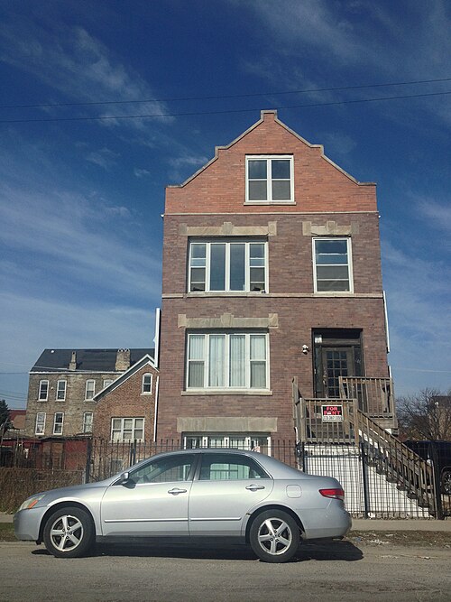 A brick two-flat with a stepped gable and a peaked dormer window, a for rent sign in the ground floor window, older buildings crowded close on either side.