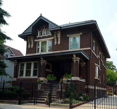 A dark red brick house with cut stone trim, a full width columned porch and a wrought iron fence along the sidewalk.