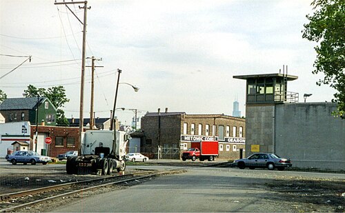 Rail tracks cross 26th Street at grade, a brick factory building lettered Metomic Corp beside the road, a small guard tower behind a chain link fence.