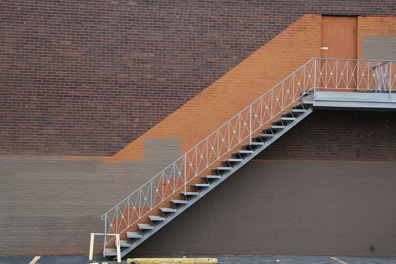 An exterior metal stair climbs a two-tone painted brick wall to a door at the second floor, a delivery ramp and parking spaces below.