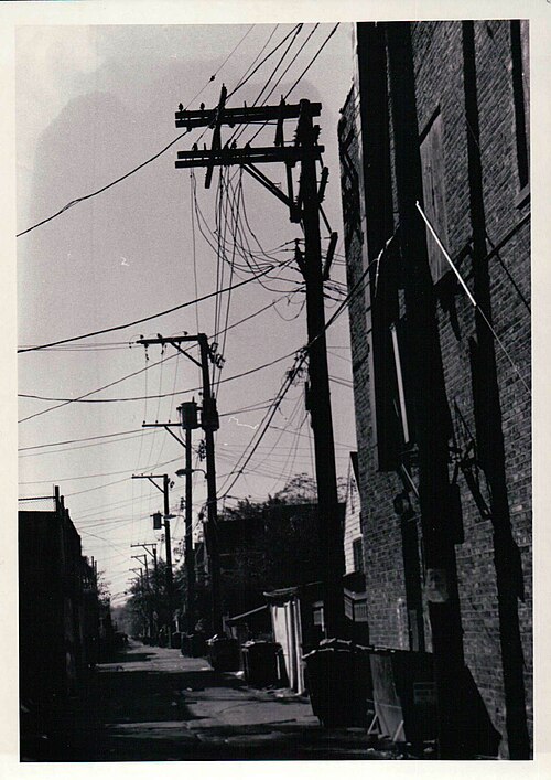 A narrow paved alley behind the 26th Street storefronts, seen in monochrome, utility poles and wires overhead and trash bins along a brick wall.