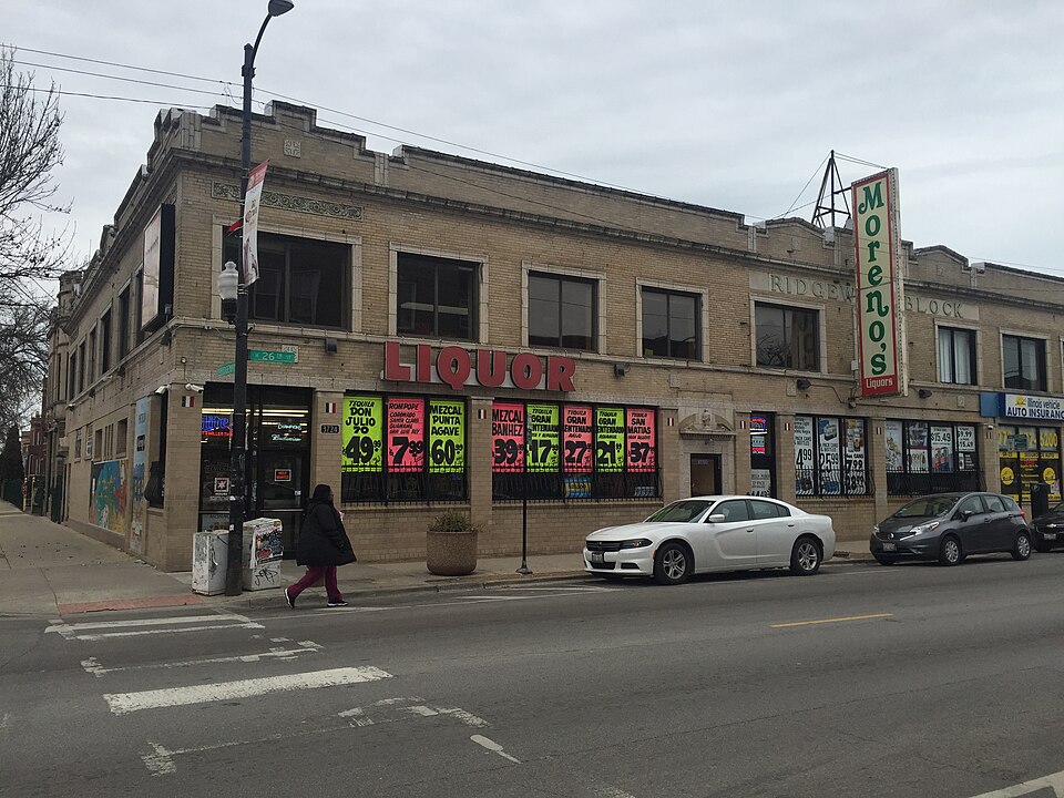 A two storey tan brick corner building with arched stone window trim and a lit sign over the sidewalk, storefront windows papered with sale posters.