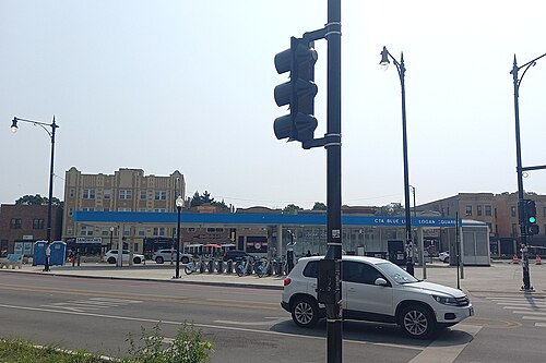 The Kedzie Avenue entrance to the Blue Line station under a blue canopy, a row of bike share docks on the sidewalk and an older stone building behind.