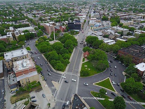 A lower, closer aerial view of the traffic circle in summer, the monument surrounded by mature trees with the crosswalks and turning lanes around it visible at street level.