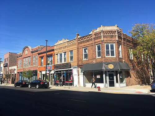 A two story ornate brick corner building on a commercial block, cornice detailing above the storefronts and a pedestrian crossing at the corner.