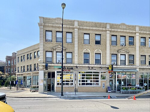 A three story tan brick building on Milwaukee Avenue with arched window surrounds, its ground floor storefronts including a bar with outdoor seating.