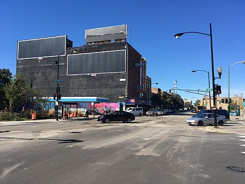 A wide intersection lined with three and four story brick buildings, blank billboards above the storefronts, streetlights and a bus shelter at the corner.